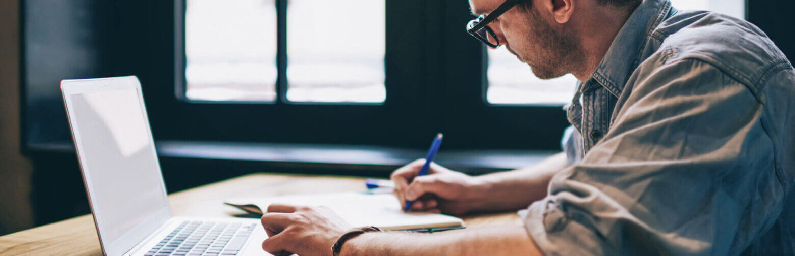 Iemand met een bril werkt aan een bureau en maakt aantekeningen naast een open laptop in een kamer met natuurlijk licht dat door een raam naar binnen schijnt.